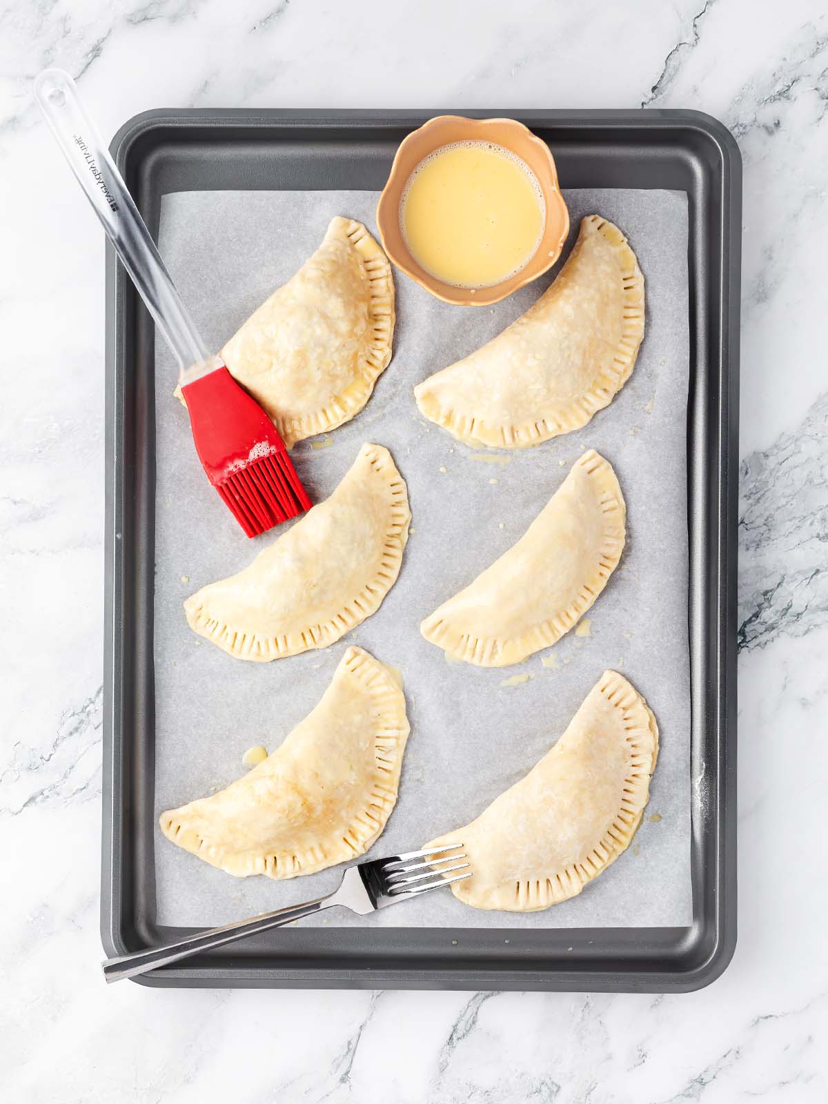 6 unbaked empanadas on a baking tray being brushed with egg wash.